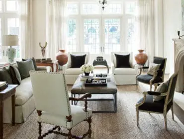 Living room with a neutral-toned sofa, upholstered armchairs, a stone fireplace, and large windows with sheer curtains.