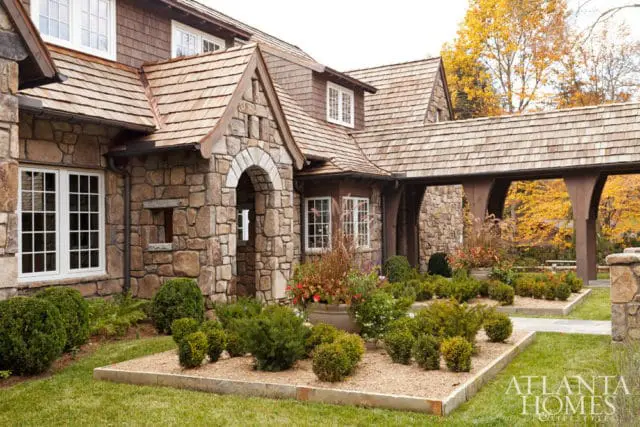 Stone and wood shingle exterior of a home with arched entryway, white grid windows, and manicured landscaping in autumn