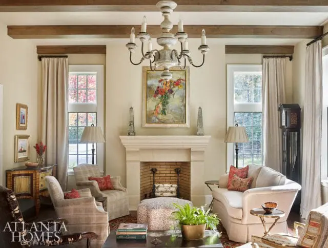 Living room with a stone fireplace, plush seating, and exposed wooden beams on the ceiling. A painting of flowers hangs above the fireplace.