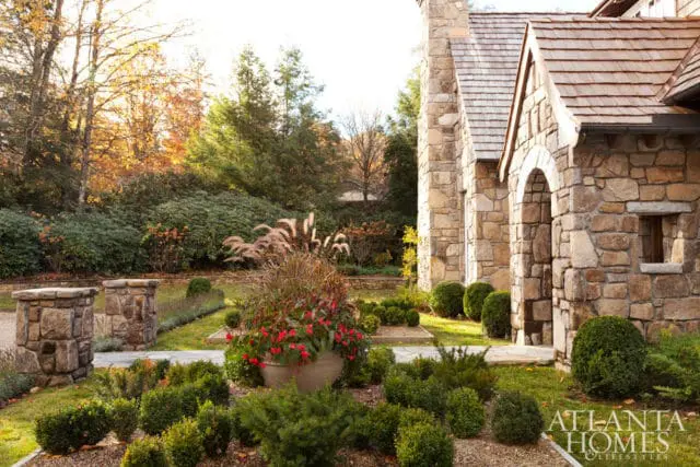Stone house with arched entryway and cedar shake roof, surrounded by manicured boxwood hedges and ornamental grasses in autumn