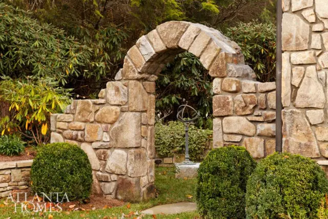 Stone archway leading to a garden with a sundial, surrounded by lush greenery and manicured boxwood shrubs.