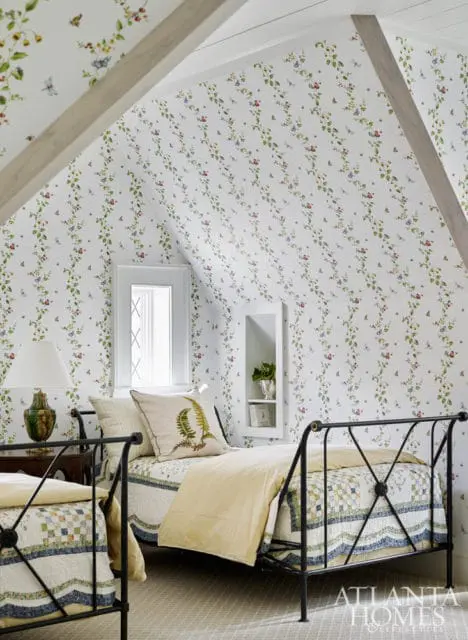 Attic bedroom with sloped ceiling featuring floral and insect wallpaper, two wrought iron beds with patchwork quilts, and a window with diamond panes.