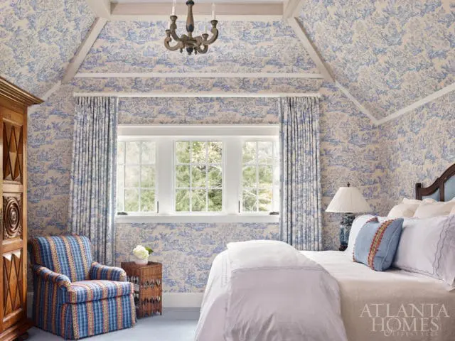 Bedroom with blue and white toile wallpaper on walls and ceiling, white trim, wooden armoire, plaid armchair, wicker side table, and a bed with white linens and blue pillows.