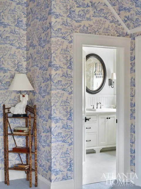 Hallway with blue and white toile wallpaper, a bamboo etagere with books and a white dog statue, and a view into a white bathroom with a round mirror.