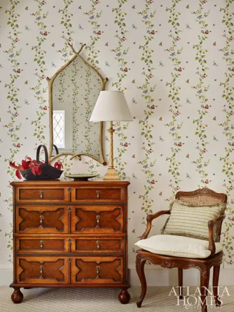 Traditional wooden chest of drawers with ornate detailing, a decorative mirror, and a cane-backed chair with cushions against floral wallpaper.