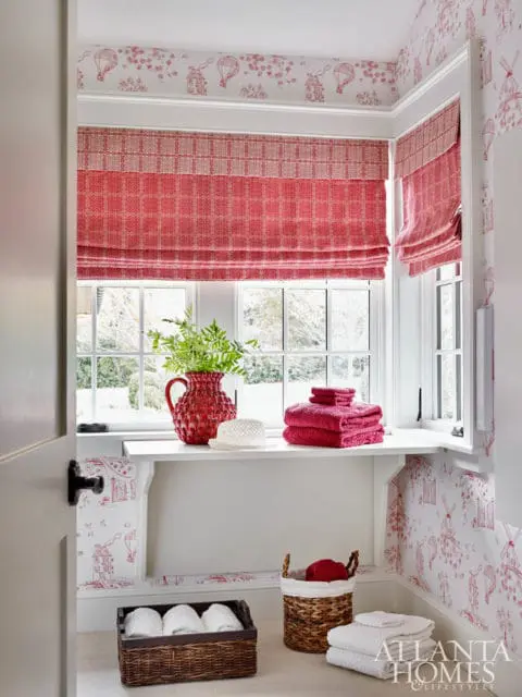 Corner of a room with red and white toile wallpaper, red patterned Roman shades, a white shelf with a red vase, folded towels, and a woven basket with towels.
