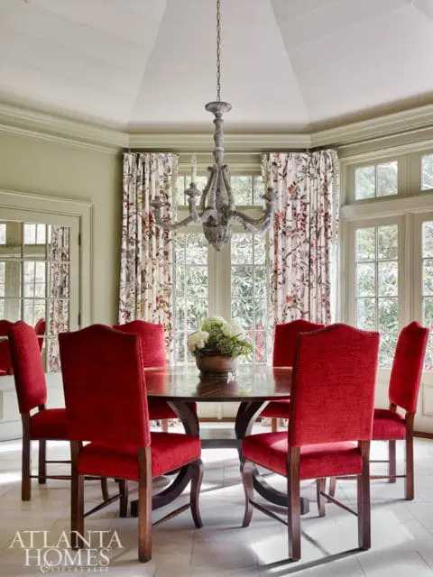 Dining room with a round wooden table surrounded by red upholstered chairs, a distressed chandelier, and floral curtains.