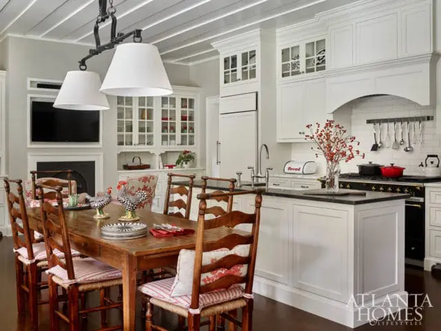 Farmhouse kitchen with white cabinetry, dark countertops, a wooden dining table with chairs, and a fireplace with a television above it.
