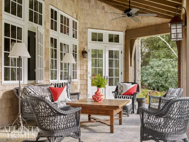 Covered porch with stone floor, shingle siding, and wood ceiling featuring a ceiling fan. Wicker furniture with patterned cushions and red accents surrounds a wooden coffee table.