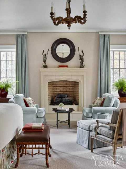 Living room with light blue upholstered chairs flanking a stone fireplace, a dark wood chandelier, and a round mirror above the mantel.