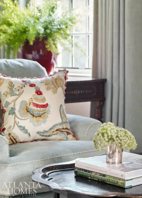 Light blue armchair with a patterned throw pillow, a silver vase of white flowers, and books on a dark tray. A large red vase with greenery sits behind the chair.