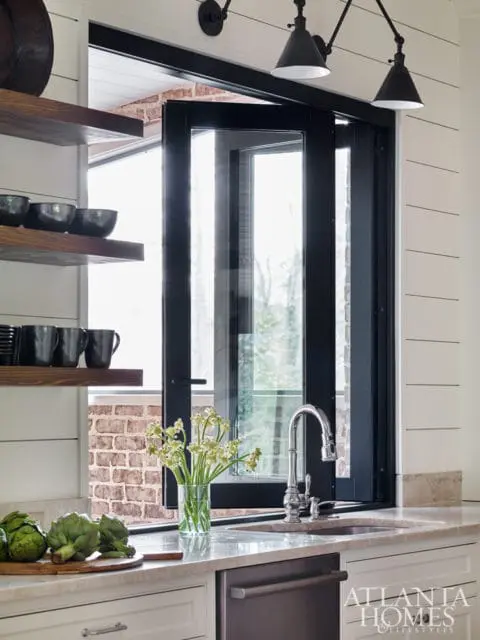 Kitchen with white shiplap walls, open wood shelving, and a large window with black framing above a sink and marble countertop.