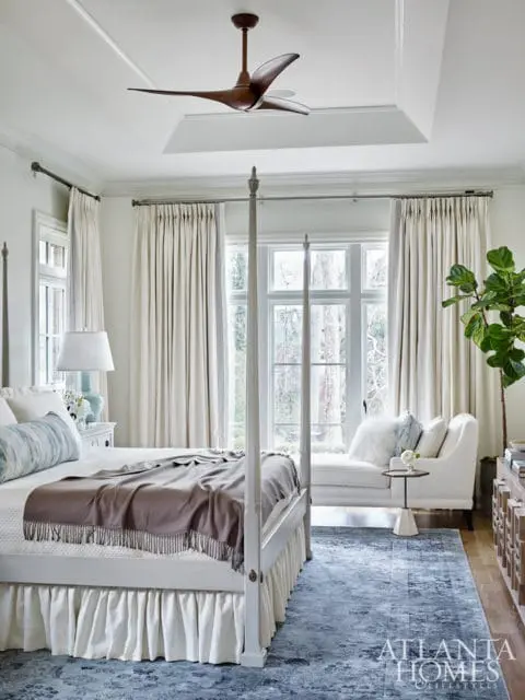 Bedroom with a white four-poster bed, draped with a taupe throw blanket, and a blue patterned rug. A white chaise lounge sits by the window, and a modern wood ceiling fan is visible.