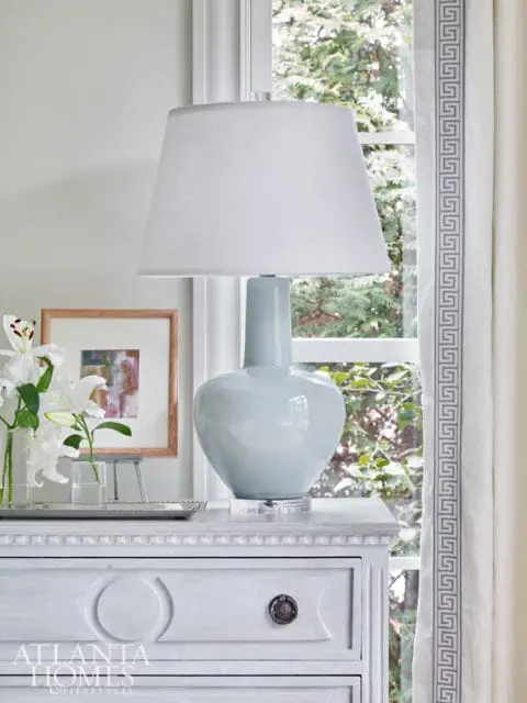 Pale blue ceramic lamp with white shade on a white dresser next to a window with Greek key patterned curtains.