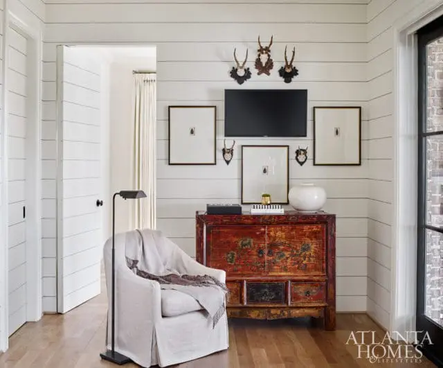 Living room with shiplap walls, a white slipcovered armchair with a throw blanket, a vintage red cabinet, framed artwork, and taxidermy mounts above a television.