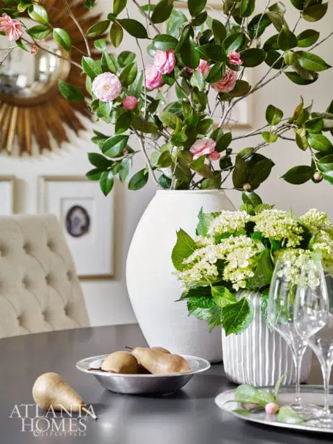 Dining table with a bowl of pears, a large white vase with pink camellias, a textured vase with white hydrangeas, and wine glasses.