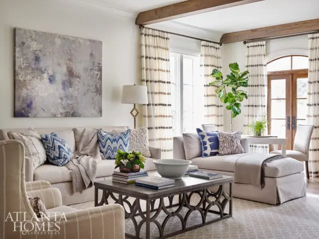 Living room with a light beige sectional sofa, abstract art, patterned curtains, and a metal coffee table with books and a bowl.