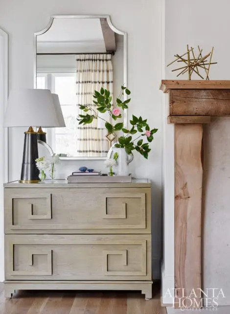 Light wood dresser with Greek key motif, topped with a metallic lamp, floral arrangement, and decorative objects. A mirror hangs above, reflecting striped curtains.