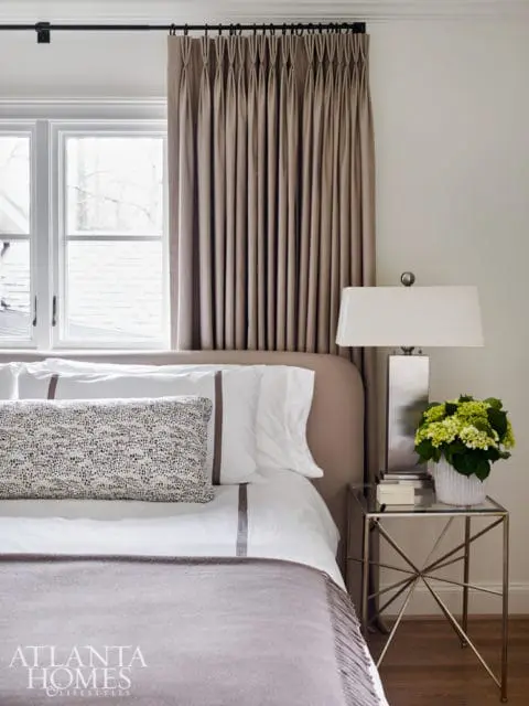 Bedroom with taupe upholstered headboard, white bedding with taupe trim, patterned accent pillow, and taupe pinch-pleat curtains. A silver bedside table holds a lamp and flowers.