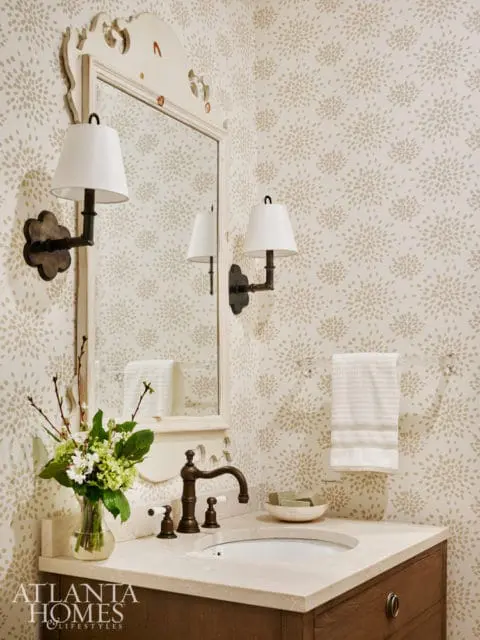 Powder room with a floral wallpaper, marble vanity, dark wood cabinet, bronze faucet, and a decorative mirror with sconces.