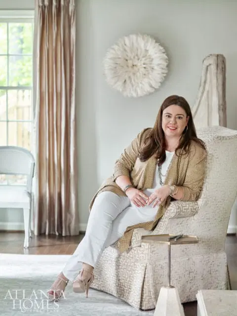 Woman in a gold patterned jacket and white pants sitting in a patterned armchair, with a large white wall decoration behind her.