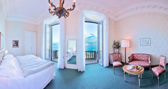 Bedroom with two beds, patterned upholstery on seating, and French doors opening to a balcony with a lake and mountain view.