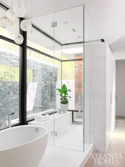 Modern white bathroom with freestanding tub, glass-enclosed shower, large windows overlooking stone exterior, and decorative pendant lights.