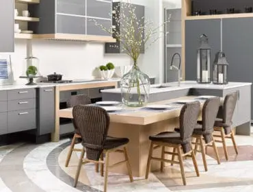 Modern gray kitchen with light wood island, wicker chairs, marble countertops, and a large glass vase with branches.