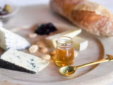Close-up of a cheese board with wedges of blue cheese and brie, almonds, a small jar of honey, and a golden spoon with a bird-shaped handle.