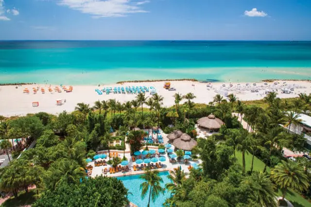 Aerial view of a tropical resort with a swimming pool, thatched-roof cabanas, and a white sand beach leading to turquoise ocean waters.