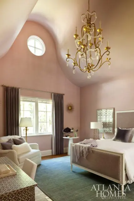 Bedroom with pink walls, vaulted ceiling, oval window, gold crystal chandelier, white armchair, and a bed with a tufted headboard.