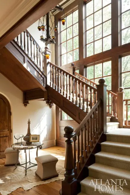 Grand wooden staircase with intricate balusters and a large window wall. A decorative chandelier hangs above.