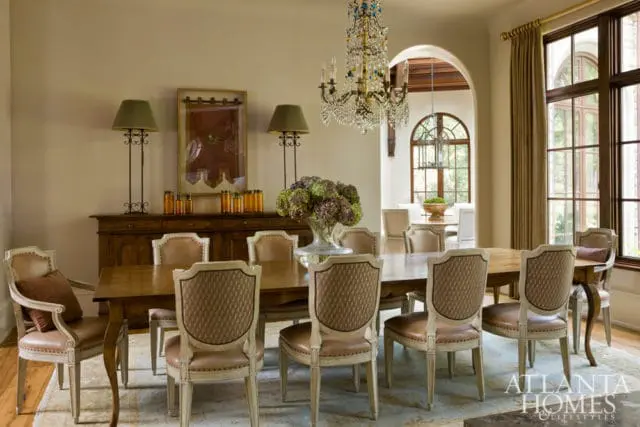 Dining room with a large wooden table surrounded by upholstered chairs, a crystal chandelier, and a buffet with decorative lamps and vases.