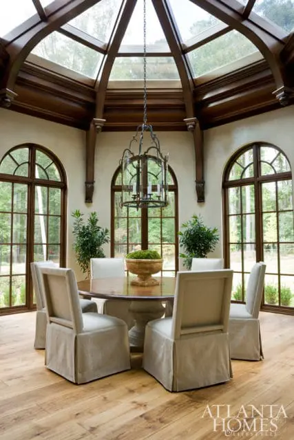 Dining room with a round wooden table, upholstered chairs, and a large glass-paneled lantern chandelier under a vaulted wood-beamed ceiling with a skylight.