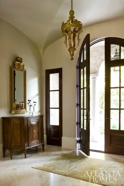 Entryway with arched double doors, a wooden cabinet with a mirror above, and a gold chandelier hanging from a vaulted ceiling.
