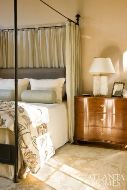 Bedroom with a four-poster bed draped in light linen curtains, a tufted headboard, and patterned bedding. A polished wood nightstand holds a lamp and a framed picture.