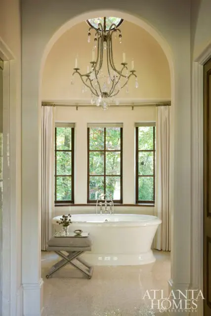 Creamy bathroom with a freestanding white tub, mosaic tile floor, and a crystal chandelier above three windows overlooking trees.