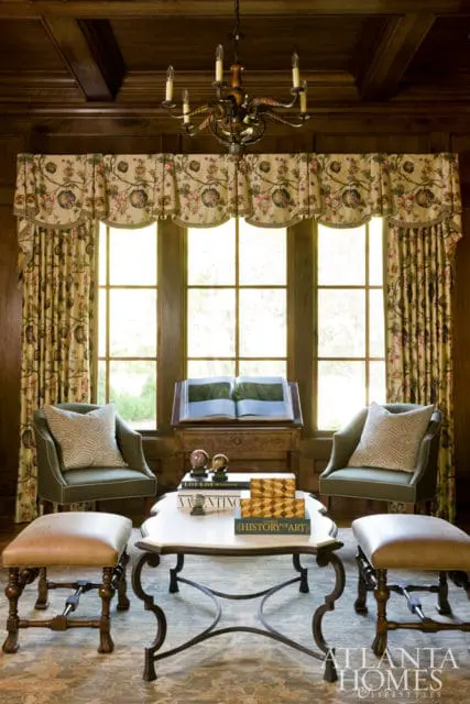 Sitting area with two green upholstered chairs, two leather ottomans, a marble-topped coffee table, and a wooden desk with an open book, beneath a floral valance and dark wood paneled ceiling.