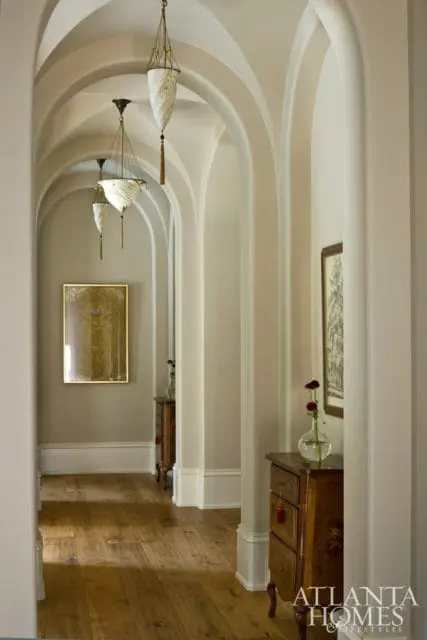 Hallway with arched openings, ornate pendant lights, wooden floors, and antique chests with artwork on the walls.