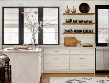 White kitchen with shiplap walls, marble countertops, and a large island with upholstered bar stools. Stainless steel dishwasher and open shelving with decorative bowls and vases are visible.