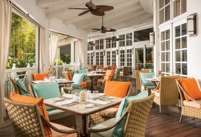 Covered outdoor dining area with tables set for lunch, featuring wicker chairs with teal and orange cushions, white curtains, and large glass doors leading inside.