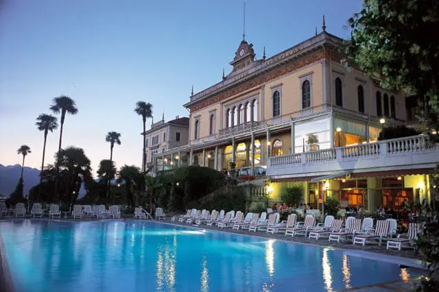 Grand Hotel Villa Serbelloni with a large swimming pool and lounge chairs in front, palm trees and mountains in the background, at dusk.