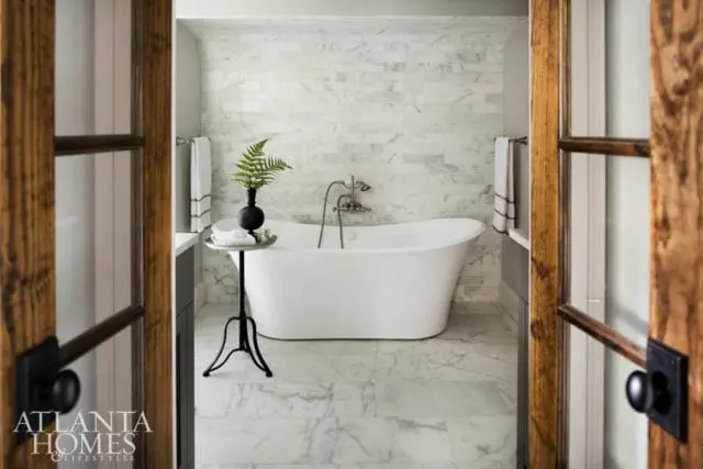 Freestanding white bathtub in a bathroom with marble tile walls and floor, dark wood door frames, and a black vase with a fern on a side table.