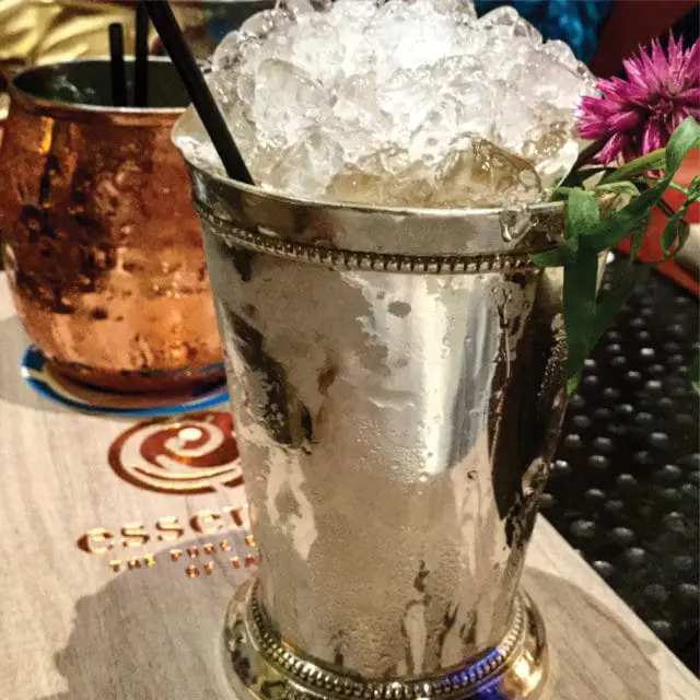 Close-up of a silver julep cup filled with crushed ice and a straw, garnished with a purple flower and greenery. A copper mug is visible in the background.