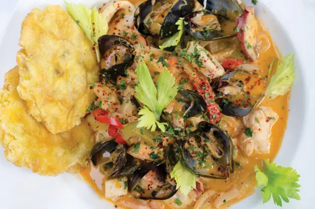 Close-up of a white bowl filled with bouillabaisse, featuring mussels, calamari, fish, and red peppers in a rich broth, garnished with parsley and celery leaves, served with two fried plantain slices.