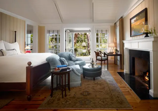 Bedroom with a dark wood four-poster bed, light blue upholstered chairs, a fireplace with a black marble hearth, and French doors opening to a view of trees and water.