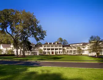 White Georgian-style mansion with multiple levels, balconies, and dormer windows, set on a manicured lawn with large oak trees under a clear blue sky.