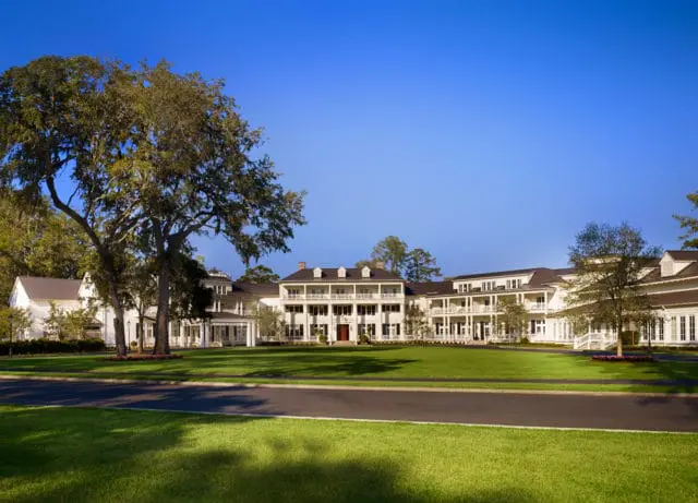 Large white building with multiple levels of balconies and dark roofs, set against a clear blue sky. A manicured lawn and mature trees surround the structure.