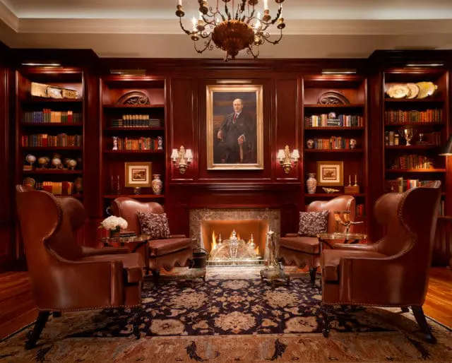 Library with dark wood built-in bookshelves filled with books, a fireplace with a portrait above, and two brown leather wingback chairs facing each other.