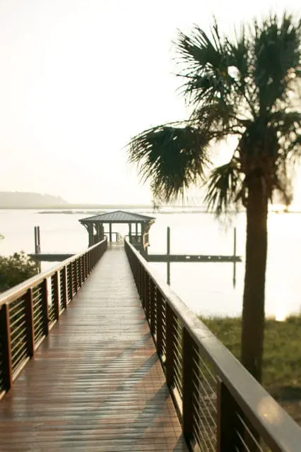 Wooden boardwalk leading to a covered dock on a calm body of water, with a palm tree in the foreground and a hazy sunrise in the background.